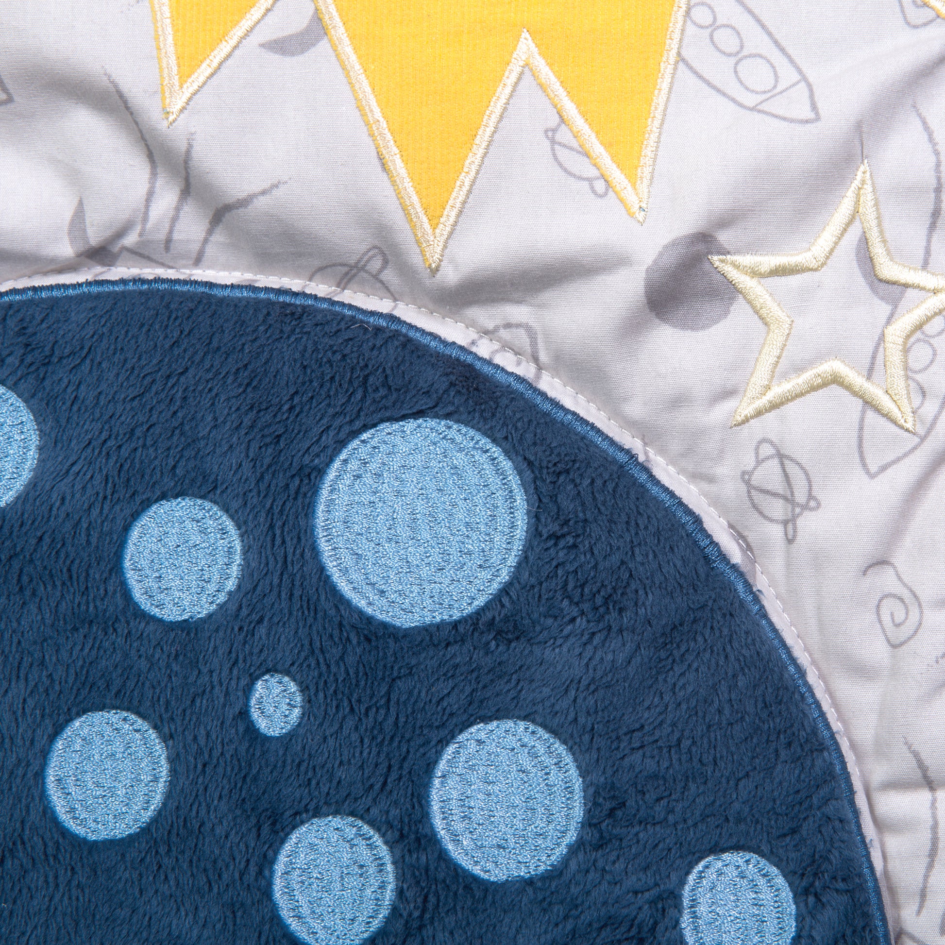Reversible 100% cotton quilt measures 35 in x 45 in and features rocket ships scatter print with planet appliques bordered by solid and star constellations in navy blue, gray, white with pops of yellow. Quilt is backed in star constellations in navy blue 