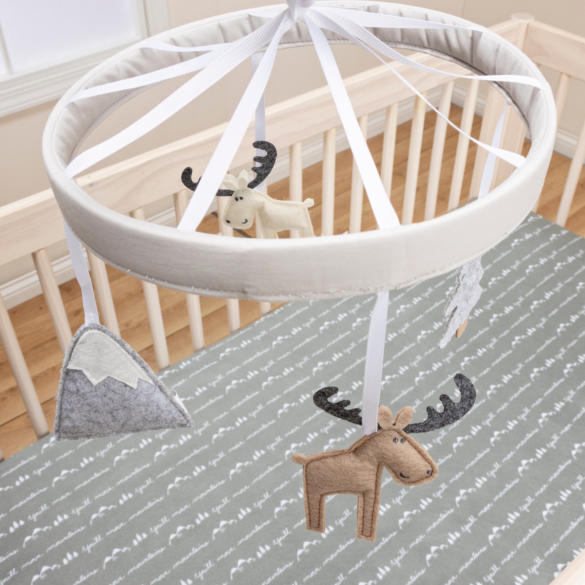 Stylized In Room Angled Image- Big Sky Musical Crib Baby Mobile- eaturing a simple solid soft gray 1-inch banded mobile ring, with a white slide cover. Two smiley moose, a mountain and a pine tree from felt are suspended from decorative white ribbons to s