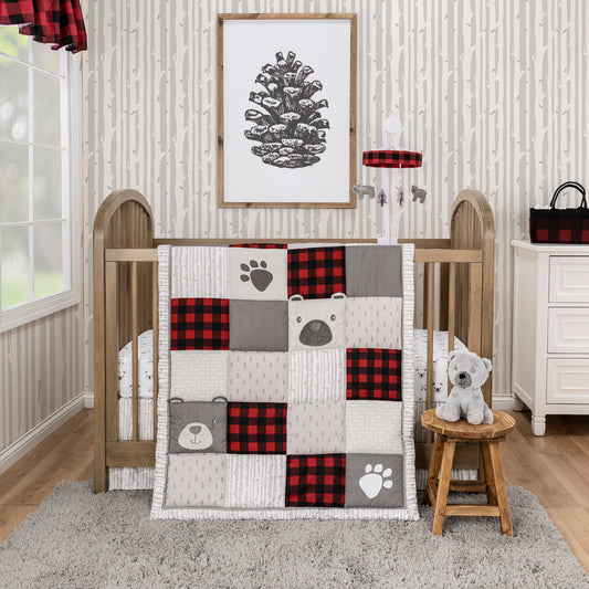 Up North 4 Piece Bedding Set in a stylized bedroom. The quilt, crib sheet & skirt sit on a tan natural wood crib, with a lumberjack musical crib mobile above it. The gray bear plush toy sits in front of the crib on a wooden stool with a gray rug and tan wood flooring below it. On the left side, a window and red and black buffalo check window valance. The walls are decorated with birch wall paper and a large pinecone wooden decor piece. 