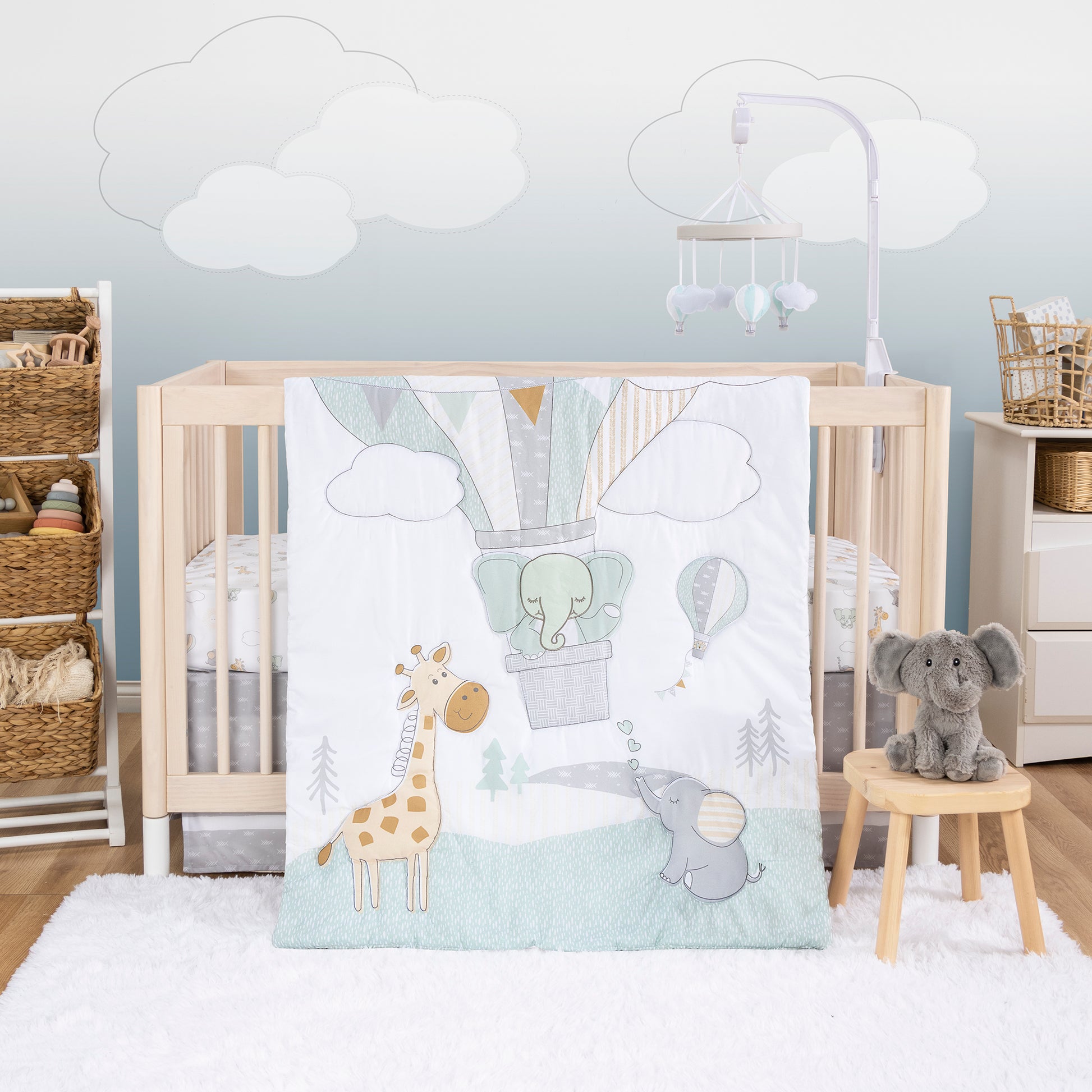 Up Up & Away 4 Piece Crib Bedding Set by Sammy & Lou® - in a stylized bedroom.  This collection features a multi patterned hot air balloon with a sweet friendly elephant surrounded by a baby elephant and a baby giraffe. With a soft muted color palette of sage green, golden orange, and gray, this design will be the perfect tranquil touch to any sweet dreamer's nursery!