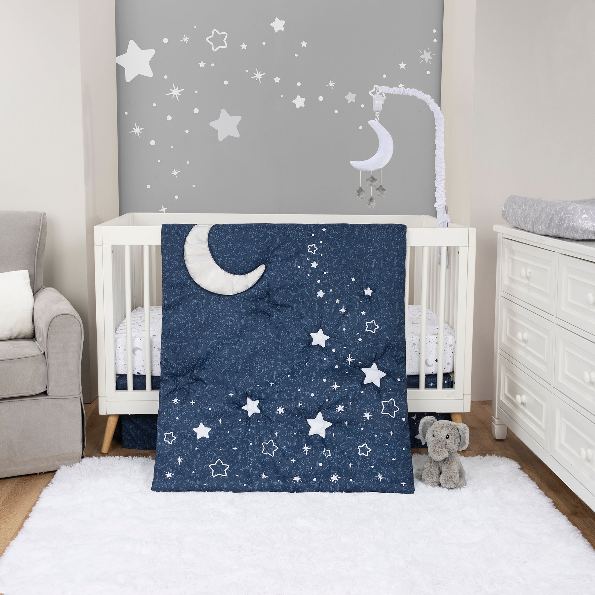 Shooting Stars 4 Piece Crib Bedding Set by Sammy & Lou® in stylized room. The wall is accented with cream pillars and a gray accent wall with scattered stars. The floor is made of a natural wooden flooring and a white rug. The nursery quilt, crib sheet and skirt, and coordinating celestial mobile (sold separtely) sit on a modern white crib with natural wooden feet. The elephant plush toy sits in front of the crib. A white dresser and gray rocking chair accents the sides of the nursery.